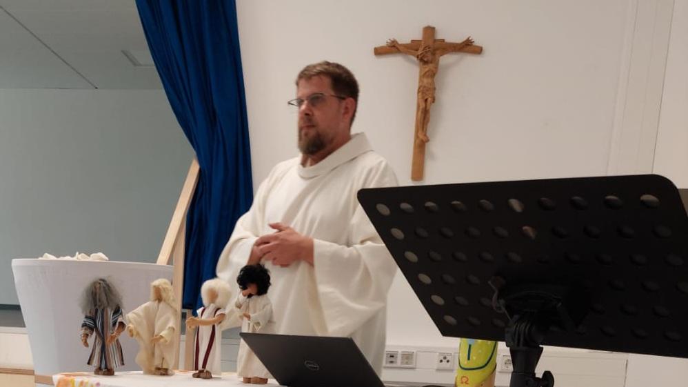 Person in liturgischer Kleidung präsentiert vor einem Tisch mit biblischen Darstellungen und Kruzifix.