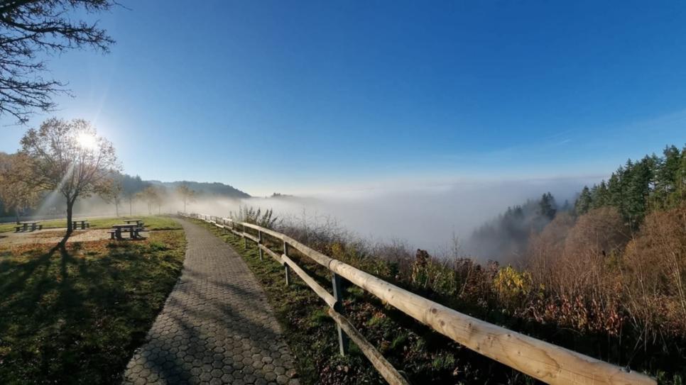 Bild mit Nebel Weg mit Holzgeländer führt zu einem Aussichtspunkt über einem nebelverhangenen Tal unter blauem Himmel.