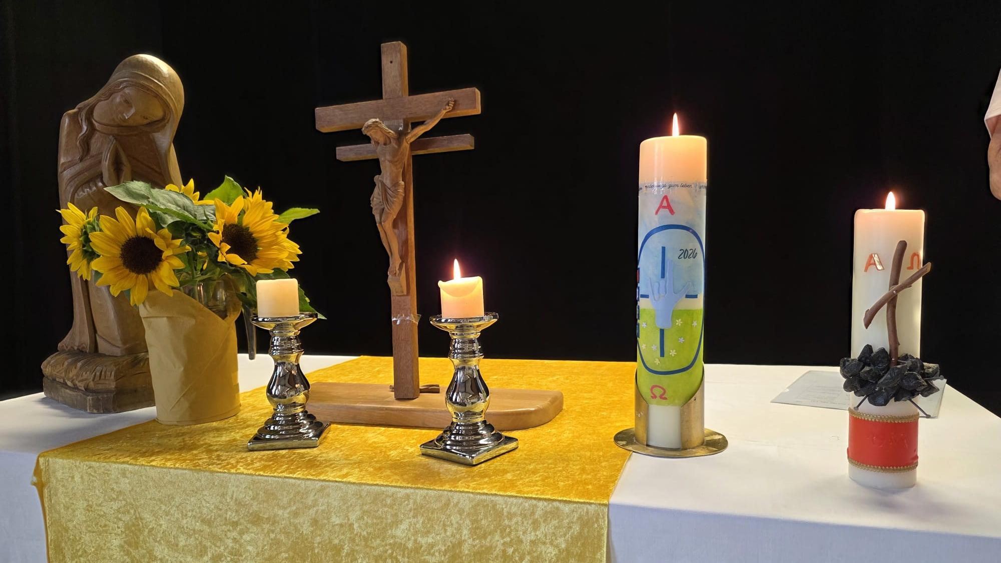 Kirchlich geschmückter Altar mit Kruzifix, Marienfigur, Sonnenblumen und brennenden Kerzen vor dunklem Hintergrund.